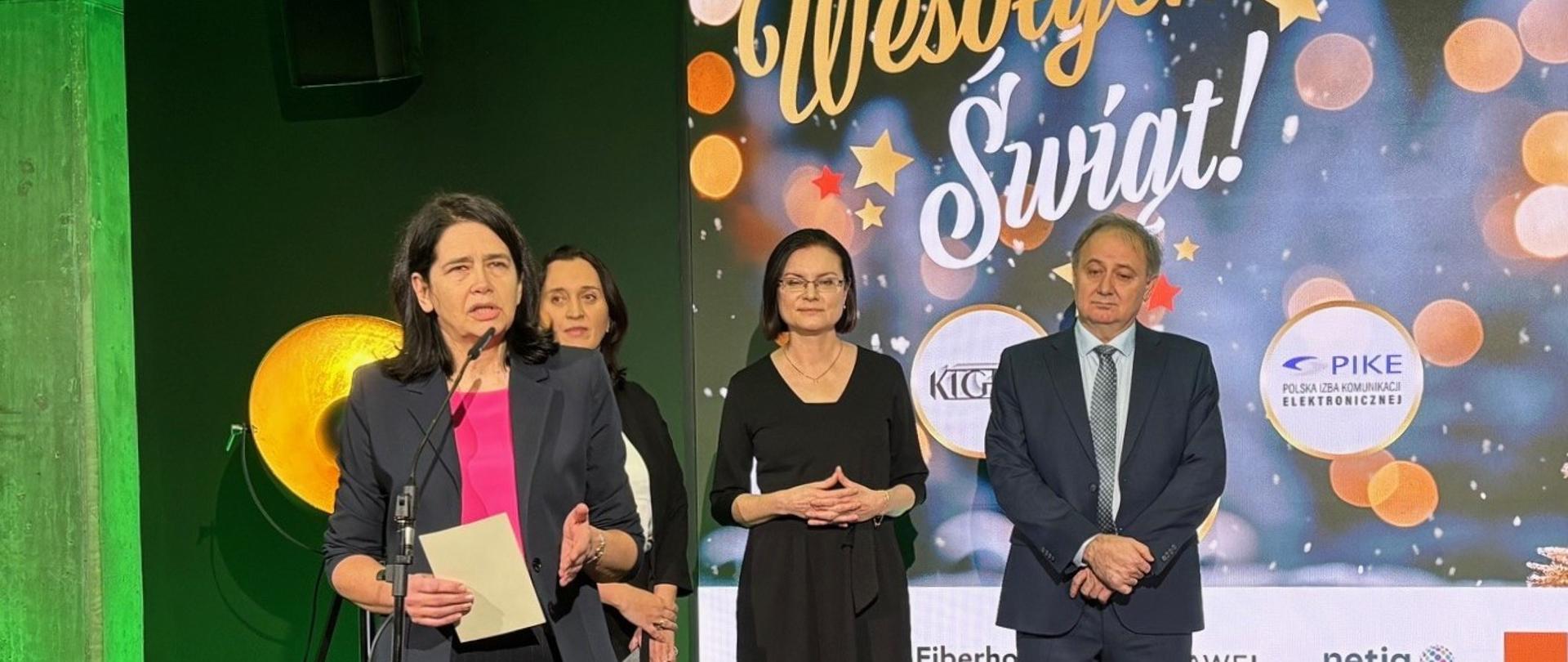 Four speakers standing on a stage during an ICT industry Christmas event, with one woman speaking at a microphone and holding notes, while three other participants stand beside her. A festive backdrop with the text “Wesołych Świąt!” and ICT industry logos is visible behind them.