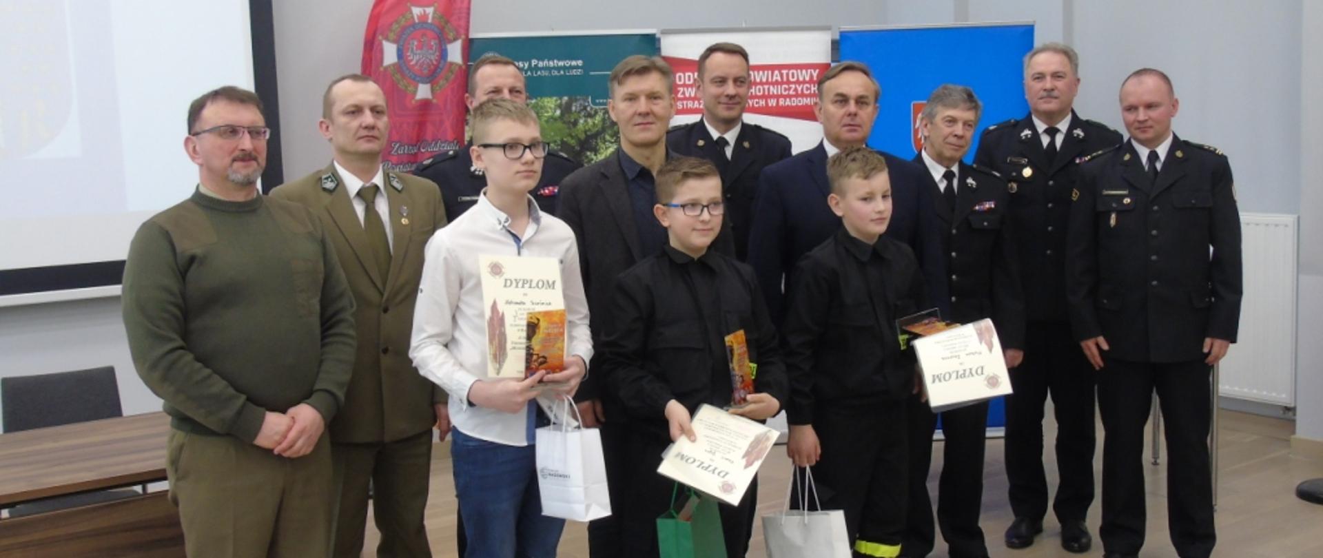 Pamiątkowe zdjęcie wykonane na zakończenie turnieju. Na zdjęciu w pierwszym rzędzie widać trzech chłopców – zwycięzców swojej kategorii wiekowej. W dłoniach trzymają pamiątkowe dyplomy oraz upominki. Za nimi w drugim rzędzie stoją przedstawiciele Lasów Państwowych, funkcjonariusze Państwowej Straży Pożarnej, Prezesi jednostek z Ochotniczych Straży Pożarnych oraz przedstawiciele lokalnych władz samorządowych. 