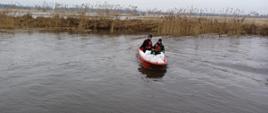 Na zdjęciu widać dwóch strażaków w czarnych strojach i kapokach, którzy płyną czerwoną łódką. Na łódce znajdują się białe worki wypełnione piaskiem. W tle widać zarośla oraz łąki, które są częściowo zalane. Jest widno.