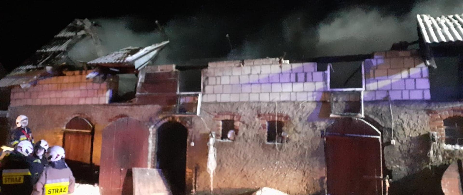 W centrum widać murowany budynek częściowo bez dachu, z którego wydobywają się kłęby dymu. Po lewej stronie widać strażaków.
