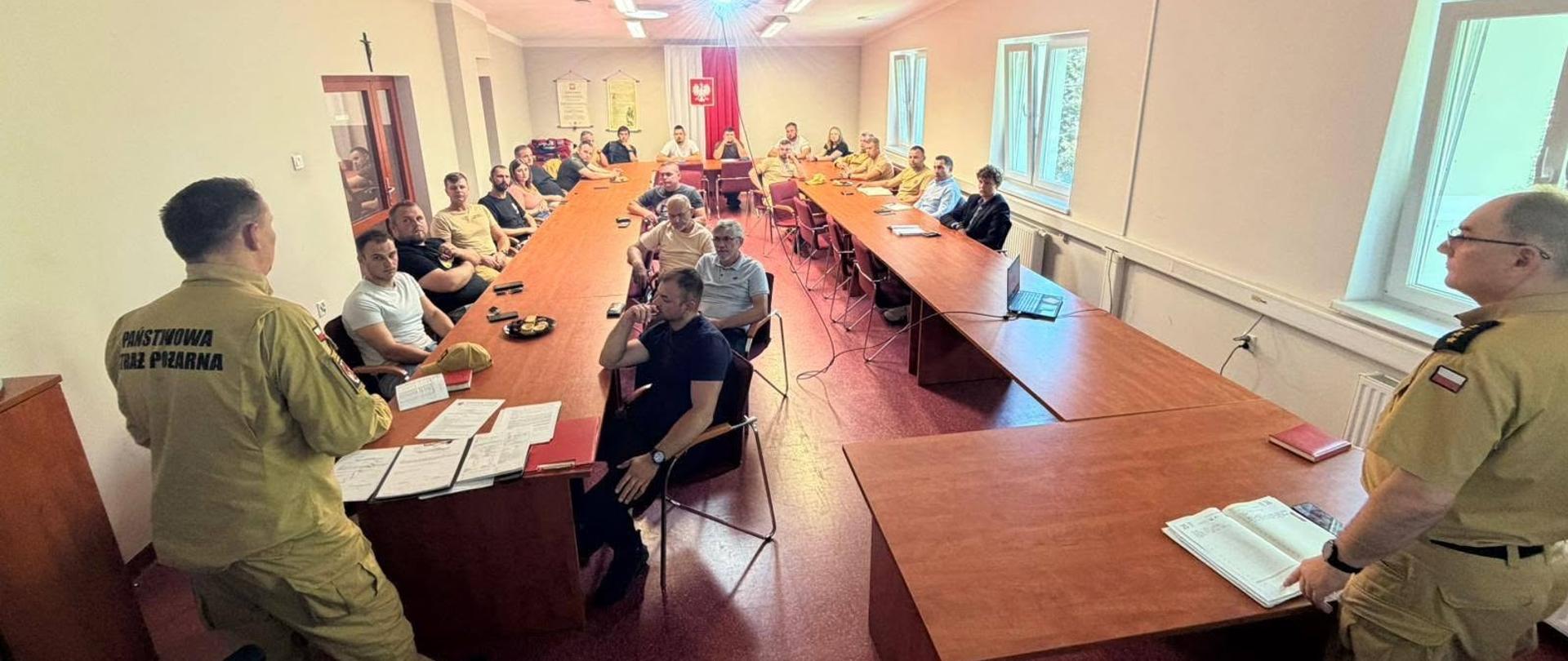 Na pierwszym planie, po lewej i po prawej stronie, stoją dwaj mężczyźni w piaskowych (beżowych) mundurach służbowych Państwowej Straży Pożarnej. Strażak po lewej stronie jest odwrócony tyłem do obiektywu, a na plecach jego bluzy widnieje wyraźny, czarny napis „PAŃSTWOWA STRAŻ POŻARNA”. Drugi mundurowy stoi po prawej stronie przodem do sali i trzyma otwarty notes lub dokument. W głębi pomieszczenia ustawione są długie stoły w kształcie litery „U” o ciemnoczerwonym lub mahoniowym kolorze. Przy stołach siedzi kilkanaście osób. Część z nich ma na sobie ubrania cywilne (koszulki polo, T-shirty), a pozostali noszą ciemniejsze stroje, co sugeruje, że spotkanie może mieć charakter szkoleniowy, informacyjny lub być odprawą z przedstawicielami innych jednostek (np. strażakami ochotnikami z OSP).