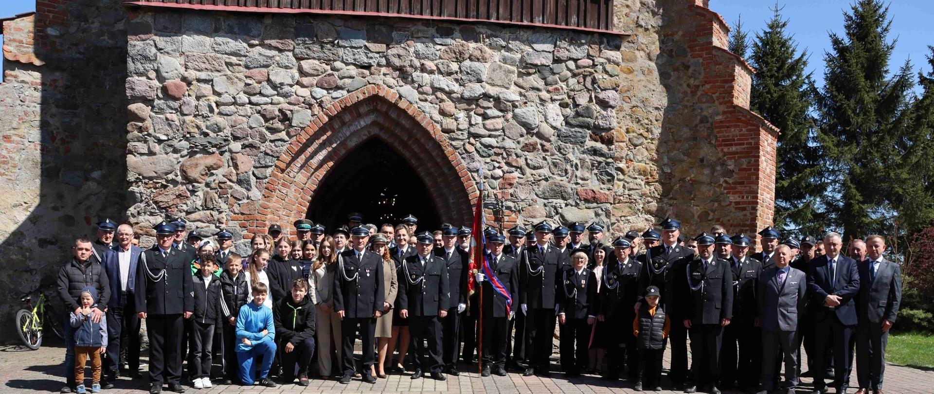 Na zdjęciu na tle zabytkowego kościoła stoi bardzo duża grupa strażaków, przedstawiciele służb oraz zaproszeni goście. Słonecznie.