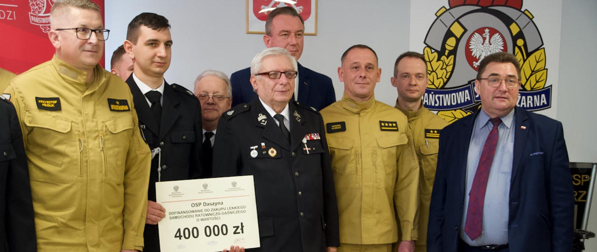 Na zdjęciu widać ośmiu mężczyzn. Dwóch ubranych jest w garnitury, trzech w mundury galowe ochotniczych straży pożarnych oraz trzech w piaskowych ubraniach strażackich Państwowej Straży Pożarnej. Jeden strażak trzyma kartkę z napisami i kwotą 400 000 złoty. W tle widać godło Polski oraz logo Państwowej Straży Pożarnej. Zdjęcie zrobione jest wewnątrz budynku.