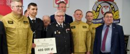 Na zdjęciu widać ośmiu mężczyzn. Dwóch ubranych jest w garnitury, trzech w mundury galowe ochotniczych straży pożarnych oraz trzech w piaskowych ubraniach strażackich Państwowej Straży Pożarnej. Jeden strażak trzyma kartkę z napisami i kwotą 400 000 złoty. W tle widać godło Polski oraz logo Państwowej Straży Pożarnej. Zdjęcie zrobione jest wewnątrz budynku.