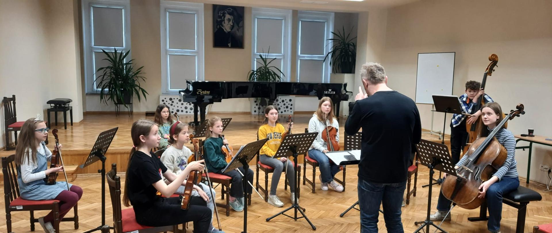 Zdjęcie przedstawiające szkolny zespół smyczkowy wraz z dyrygentem podczas warsztatów.