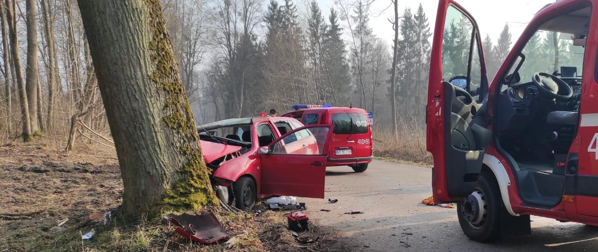Z lewej strony drzewo a przy min rozbite czerwone auto, otwarte drzwi, uszkodzona maska i wybite szyby. Z prawej strony i w oddali stoi na drodze samochód strażacki.