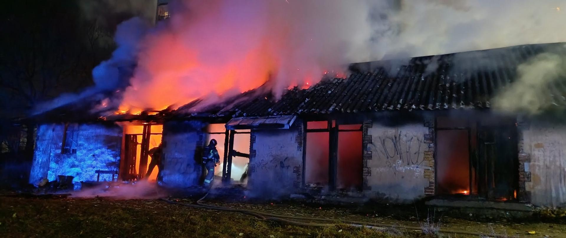 Budynek gospodarczy w płomieniach przy nim strażacy, pora nocna