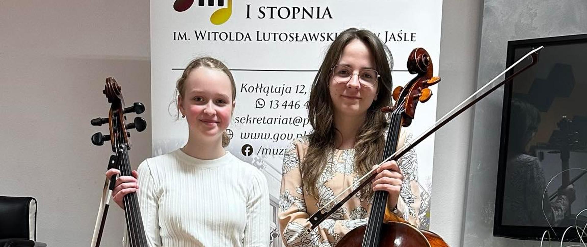 Pani wiolonczelistka oraz uczennica stoją w Sali kameralnej PSM trzymając wiolonczele. W tle rollup PSM. Ogólna kolorystyka szaro-brązowa.