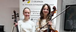 Pani wiolonczelistka oraz uczennica stoją w Sali kameralnej PSM trzymając wiolonczele. W tle rollup PSM. Ogólna kolorystyka szaro-brązowa.
