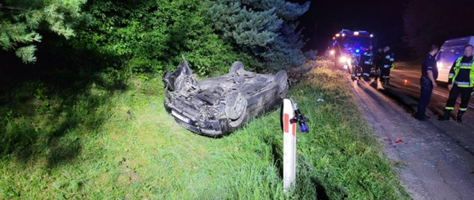 Zdjęcie przedstawia samochód osobowy, który leży na dachu w przydrożnym rowie. Przy samochodzie , na jezdni stoją funkcjonariusze i pojazdy służb ratowniczych. 
