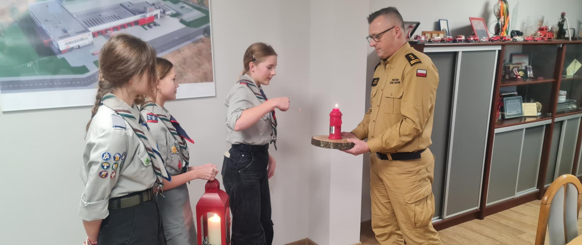 Jasne pomieszczenie. Po lewej trzy harcerki w mundurkach stojące w rzędzie z lampionem. Po prawej strażak w piaskowym mundurze z czerwoną świecą na podstawce w trakcie przekazywania ognia.
