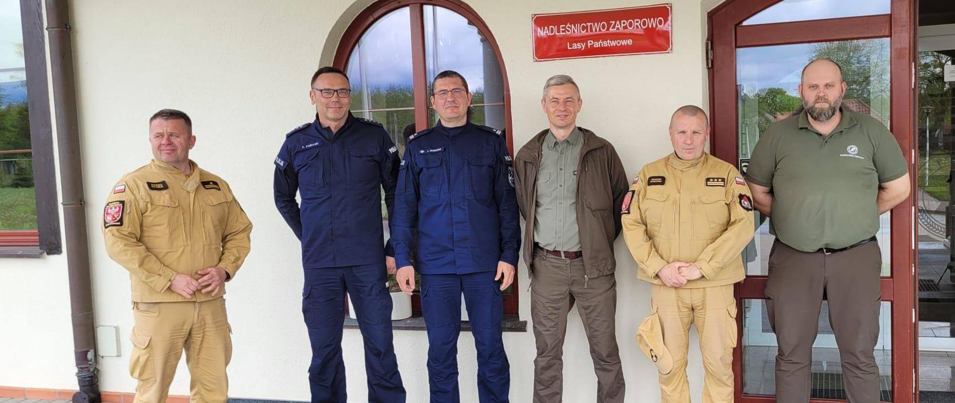 Na zdjęciu dwóch przedstawicieli lasów państwowych, dwóch policjantów i dwóch strażaków w jasnych mundurach. Stoją przy wejściu do budynku nadleśnictwa.