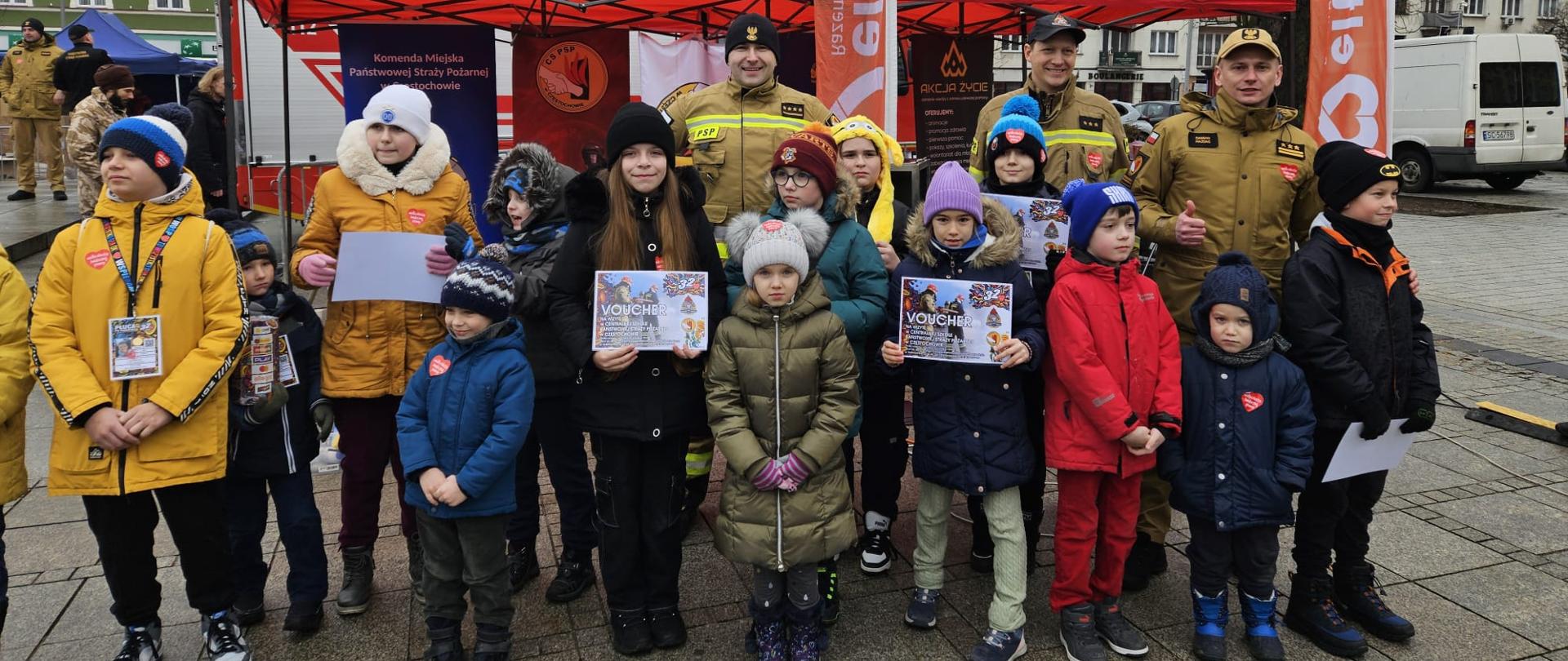 Pamiątkowe zdjęcie grupy dzieci stojącej przed stoiskiem CS PSP na placu im. dr W. Biegańskiego podczas 32. finału WOŚP, wraz z dziećmi stoi p.o. Komendant Centralnej Szkoły PSP oraz trzej inni funkcjonariusze CS PSP