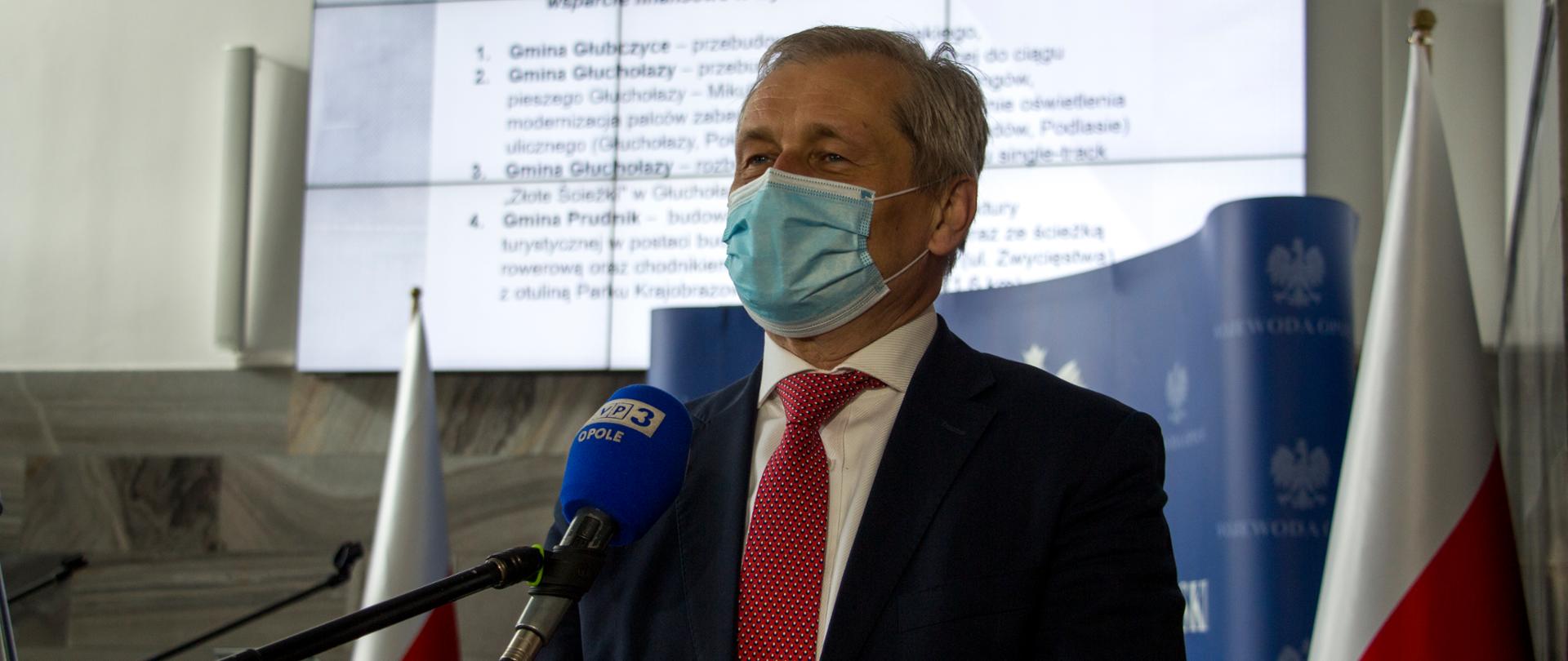 Wojewoda opolski przemawiający do dziennikarzy poprzez stojący na statywie mikrofon. Za nim w tle dwie biało-czerwone flagi, ścianka granatowa z logo OUW oraz - za jego plecami - duży monitor multimedialny z wyświetloną prezentacją, która była emitowana podczas konferencji prasowej. Wojewoda ubrany jest w ciemną marynarkę, czerwony krawat. Na twarzy ma maseczkę ochronną, patrzy w kierunku dziennikarza.