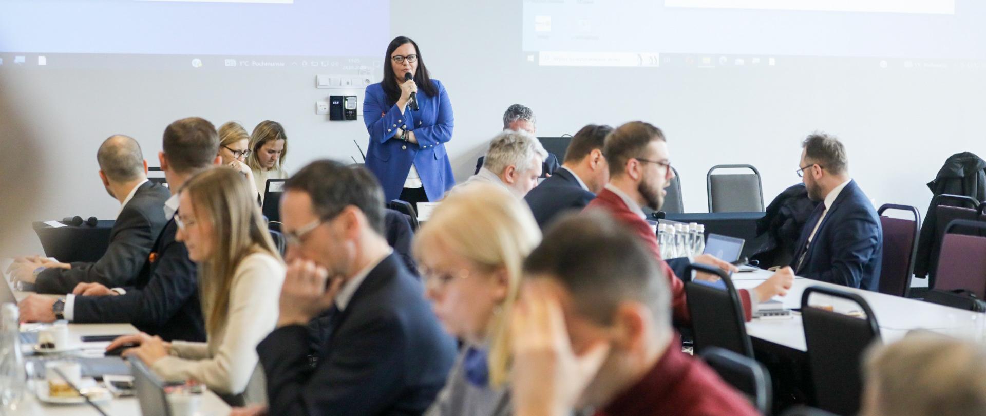 W sali konferencyjnej wiceminister Małgorzata Jarosińska-Jedynak stoi z mikrofonem. Przed nią siedzą przy stołach osoby.
