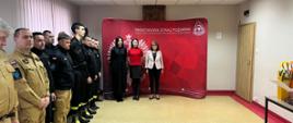 Strażacy na świetlicy, z lewej strony strażacy stoją w szyku. W tle 3 kobiety w cywilnym ubraniu na czerwonym tle z logiem psp. 