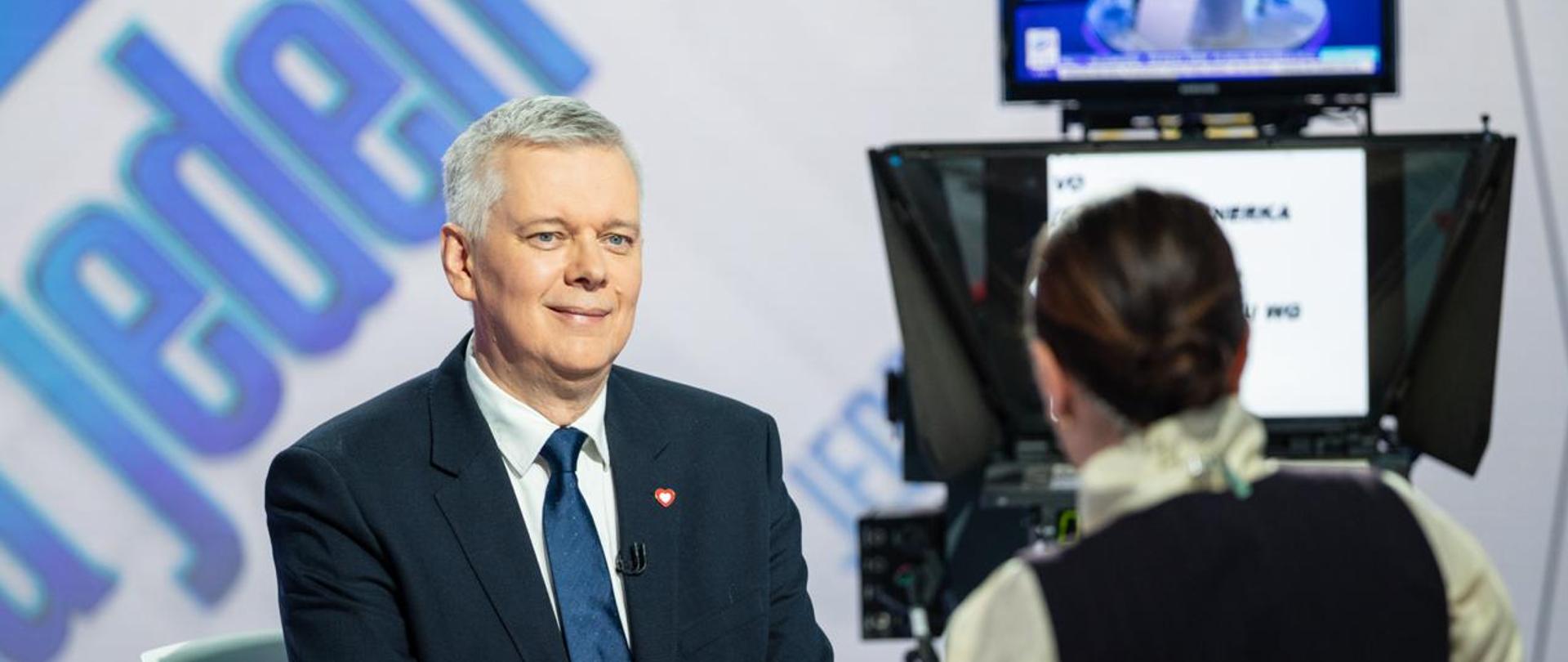 Na zdjęciu widać wnętrze studia TVN24. Przy stole siedzą dwie osoby: minister Tomasz Siemoniak (po lewej) oraz redaktor Agata Adamek (po prawej, siedzi tyłem do obiektywu). Na stole szklanka z wodą. Po prawej stronie widać prompter oraz monitor. W tle niebieski napis na białym tle - nazwa programu "Jeden na jeden". 