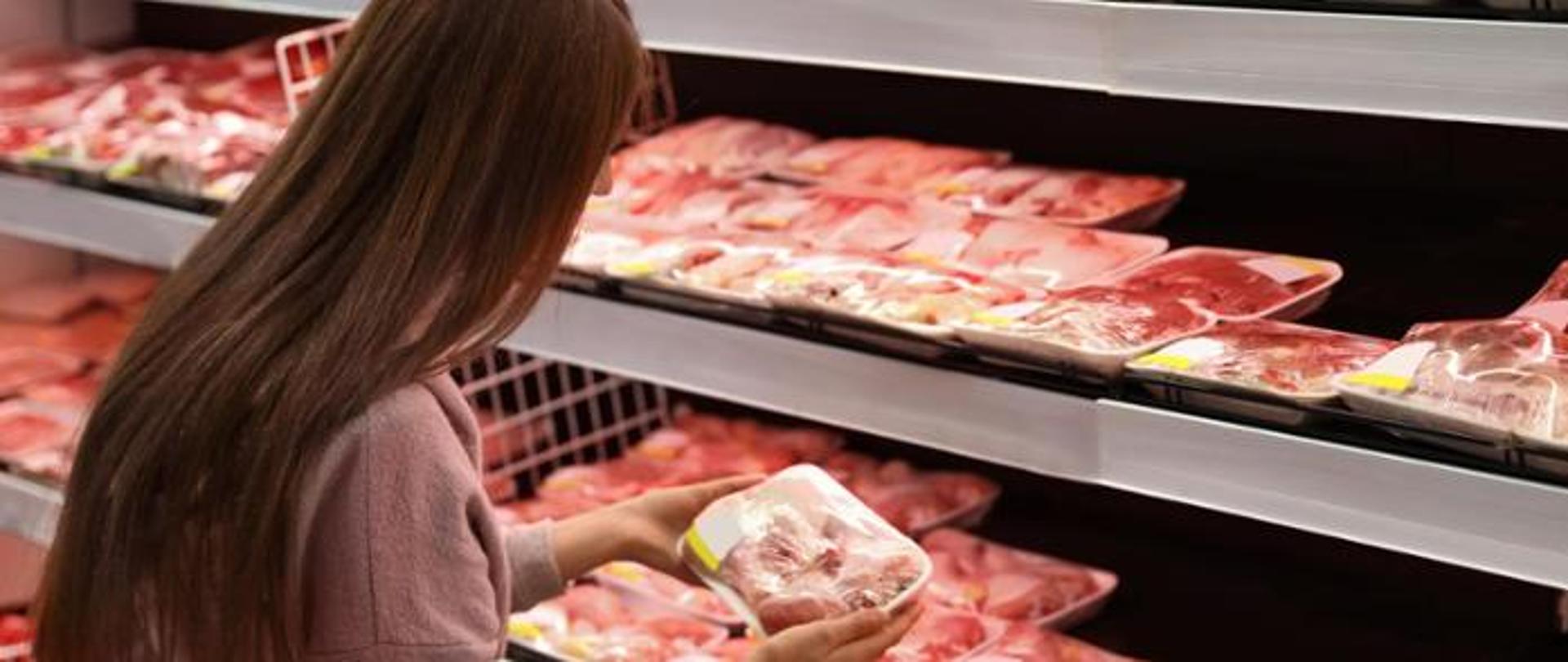 kobieta stoi przodem do półek z mięsem w sklepie i wybiera opakowanie mięsa do zakupu