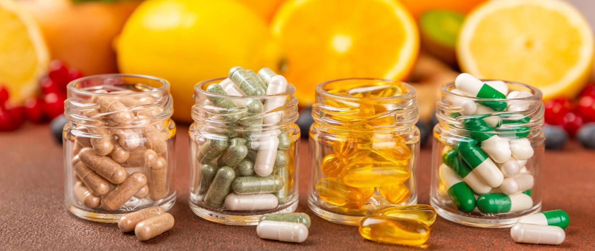Grafika przedstawia 4 słoiczki. W każdym znajduje się kilkanaście sztuk różnych kapsułek, wszystko na tle owoców.