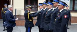 Uroczystość Dnia Strażaka: nadbrygadier w ciemnogranatowym mundurze wyjściowym z lampasami na spodniach przypina medal strażakowi stojącemu w szeregu. Obok nich inni strażacy w mundurach wyjściowych z niebieskimi teczkami w dłoniach. Po lewej stronie mężczyzna w garniturze i strażak w mundurze wyjściowym obserwują ceremonię.