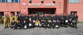 Grupa strażaków w umundurowaniu pozuje do wspólnego zdjęcia przed budynkiem KP PSP w Turku, trzymając w rękach zaświadczenia o ukończeniu szkolenia. 