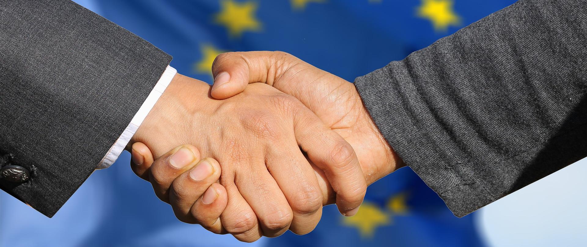witające się dłonie, za nimi flaga Unii Europejskiej