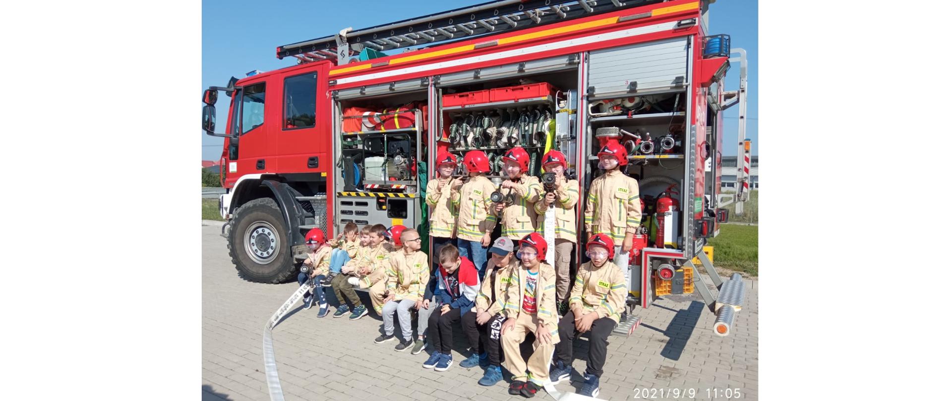 Zdjęcie przedstawia dzieci z Zespołu Placówek Edukacyjno–Wychowawczych w Gołdapi ubrane w ubrania strażackie i hełmy na tle pojazdu pożarniczego z widocznym sprzętem ratowniczym