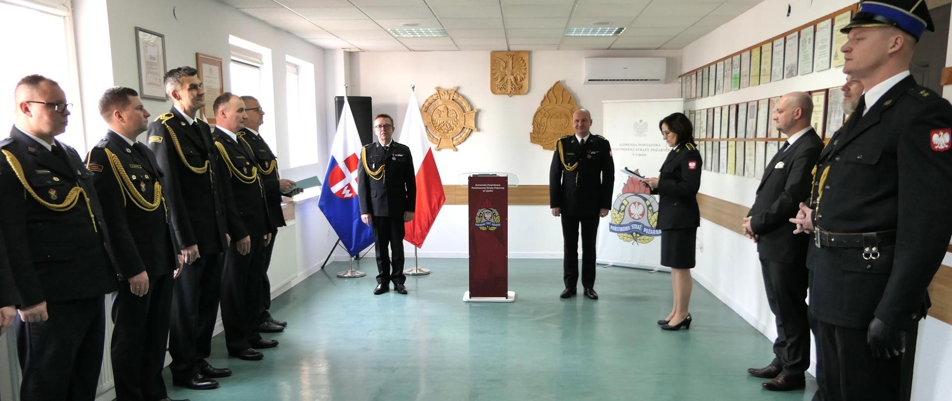 Szerokie ujęcie sali, na której odbywa się zbiórka. Strażacy ustawieni są w dwóch szeregach wzdłuż ścian bocznych. Na środku, między nimi, stoi kobieta w mundurze strażackim, która odczytuje treść z trzymanego arkusza papieru. W głębi sali, przy flagach i mównicy, stoją dwaj dowódcy. Pomieszczenie jest dobrze oświetlone światłem dziennym wpadającym przez okna po lewej stronie.