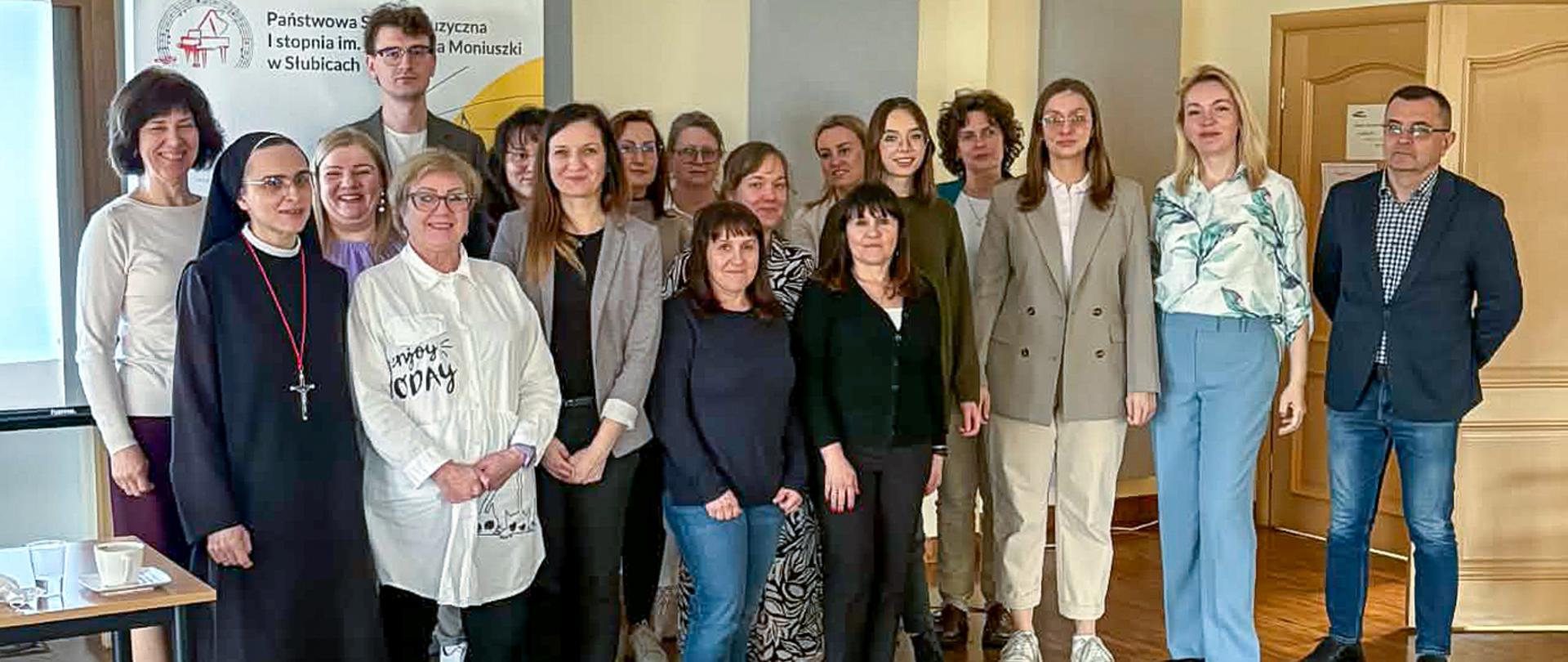 Na zdjęciu grupa kilkunastu dorosłych osób stoi w auli szkolnej.