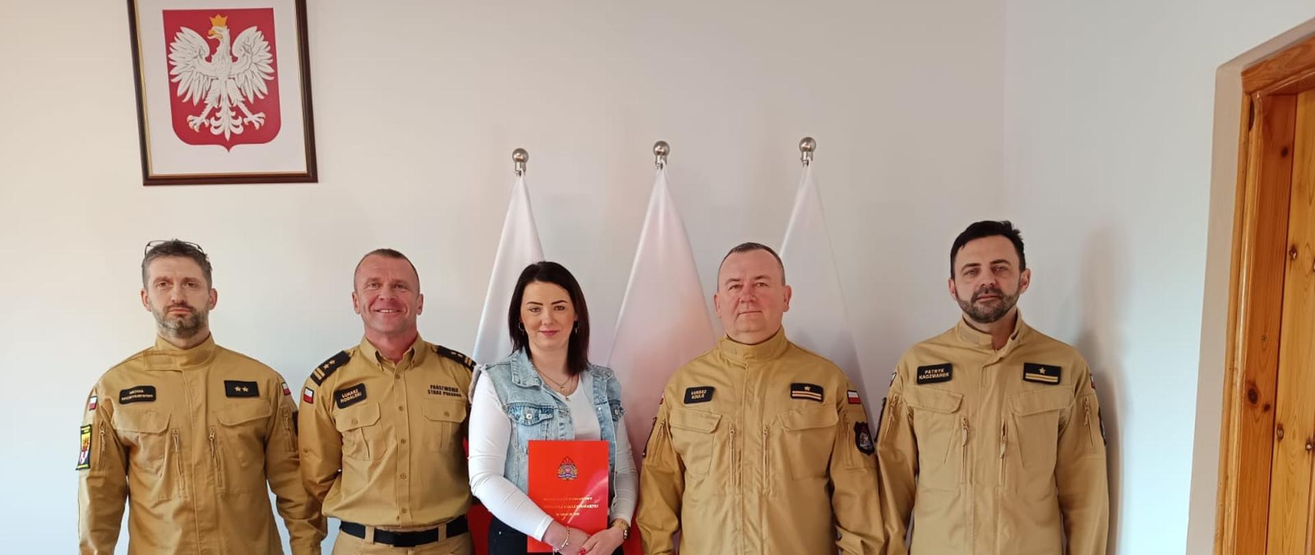 Pracownica Służby Cywilnej w towarzystwie z-cy komendanta oraz dowództwa JRG Mogilno i księgowego mogileńskiej komendy. W tle flagi Polski i wiszące na ścianie godło Polski.