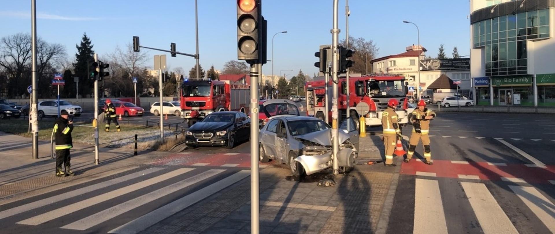 Na zdjęciu widoczne są dwa samochody biorące udział w wypadku. Jeden z samochodów uderzył w słup sygnalizacji świetlnej. Obok niego dwóch ratowników przygotowujących sprzęt. Za nimi drugi samochód biorący dział w zdarzeniu. 