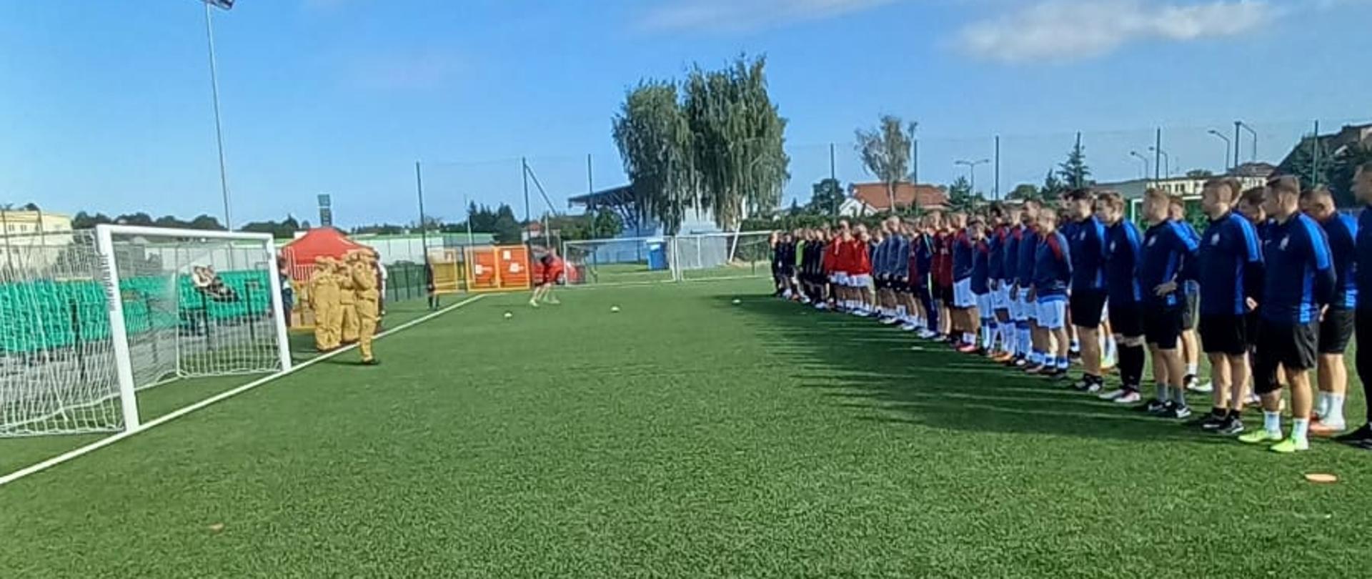 Zdjęcie przedstawia zbiórkę na zielonym boisku piłkarskim ze sztuczną nawierzchnią pod bezchmurnym, niebieskim niebem. Po prawej stronie kadru stoi długi szereg mężczyzn w sportowych strojach (niebieskie koszulki i czarne spodenki). Po lewej stronie, w oddali przy bramce, widać mniejszą grupę osób w jasnobrązowych mundurach.