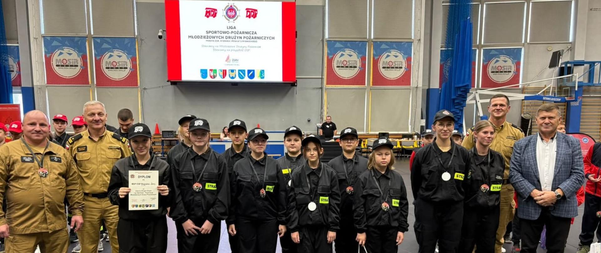 grupa młodzieży w czarnych ubraniach strażackich z żółtymi odblaskami i czarnymi czapkami stoi w szeregu na hali sportowej. Jedna osoba trzyma dyplom, inna torbę z upominkami, kilka osób ma medale na szyi. Z lewej i prawej strony stoją dorośli w mundurach, a w tle widoczny jest ekran z napisem "Liga Sportowo-Pożarnicza Młodzieżowych Drużyn Pożarniczych" oraz żółte trybuny.