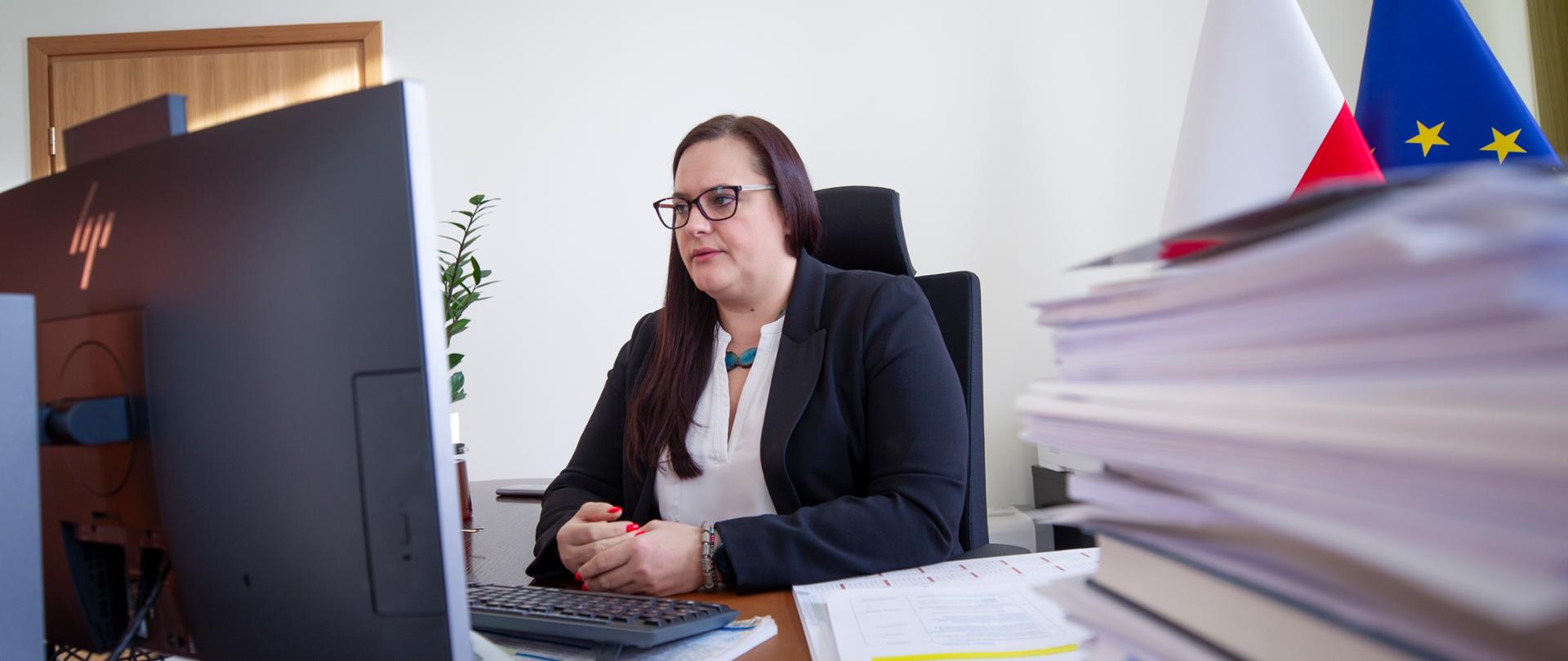 Wiceminister M. Jarosińska-Jedynak w gabinecie siedzi przy biurku, na którym stoi monitor. Obok niego widać stos kartek. Z tyłu dwie flagi unijna i polski.