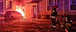 Na zdjęciu widoczny jest budynek przy którym ustawiony jest palący się samochód, pracujący strażacy.