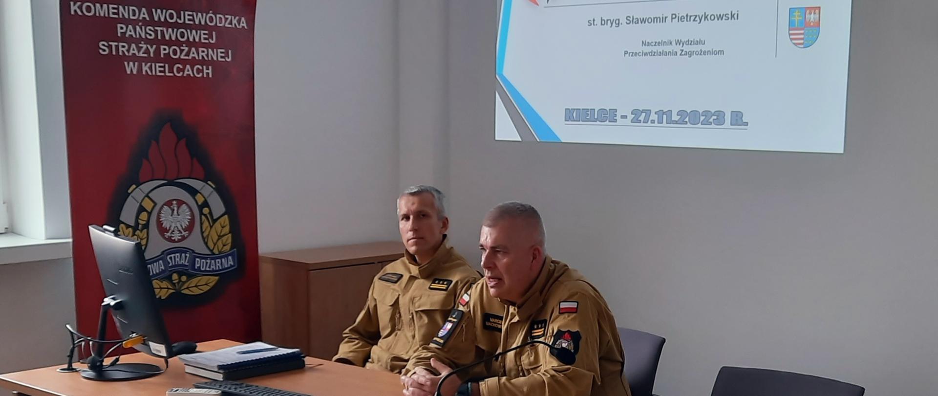 Na zdjęciu widzimy strażaków siedzących w mundurach piaskowych za stołem podczas narady służbowej. 