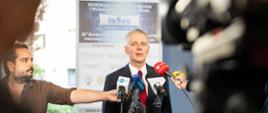 Na zdjęciu minister Tomasz Siemoniak podczas briefingu dla mediów na konferencji InSec 2026. Przed ministrem mikrofony na statywach. Na gąbkach nazwy redakcji m.in.: TVP INFO, TVN24, Polsat News, RMF FM, TOK FM, Polskie Radio. Za ministrem widać biały rollap z nazwą konferencji InSec oraz informacjami o patronach i sponsorach wydarzenia. Po lewej stronie w brązowej koszuli widać redaktora Tomasza Fenske z Radia TOK FM. W tle budynki i zieleń. Z prawej strony zdjęcia widać fragment czarnej kamery.