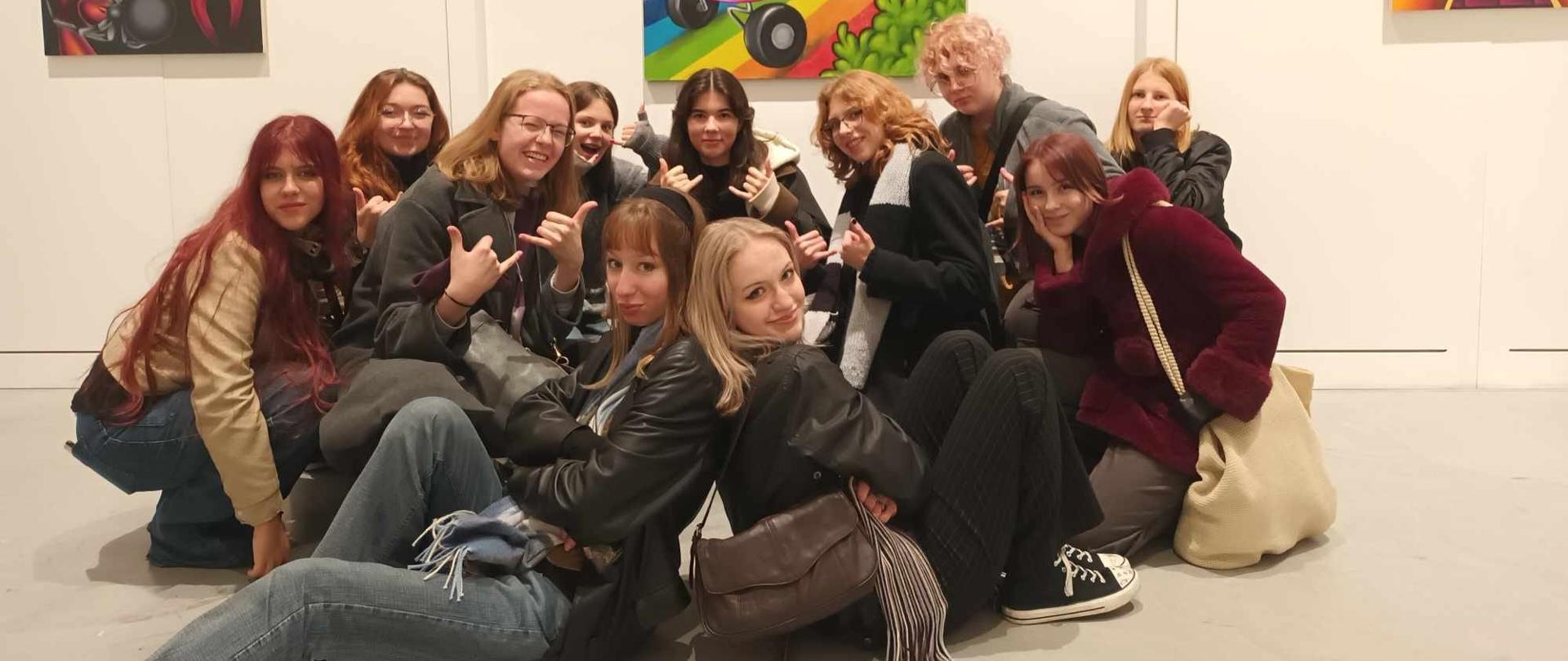Grupa uczennic w pozycji przysiadu, półleżącej, klęku pozują do zdjęcia w jednej z sal galerii warszawskiej. Dziewczęta są uśmiechnięte i zadowolone.