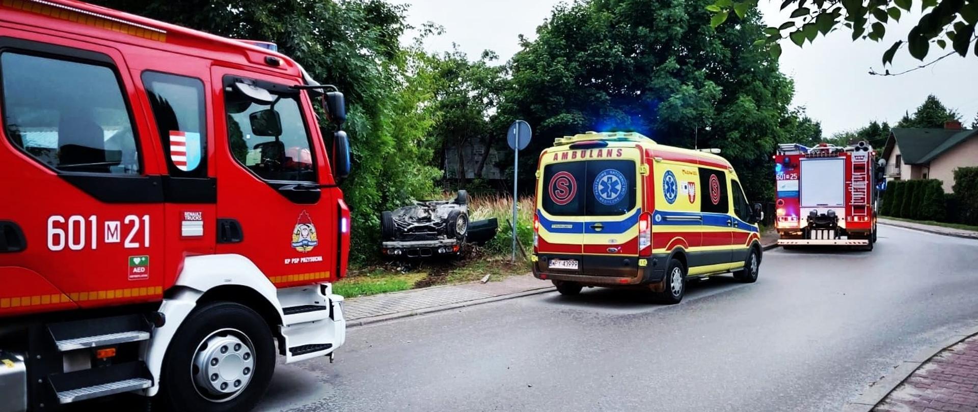 Zdjęcie przedstawia scenę wypadku drogowego, gdzie na poboczu drogi leży przewrócony samochód, widoczny w oddali w rowie lub krzakach. Na pierwszym planie po lewej stronie znajduje się duży, czerwony wóz strażacki z numerem "601-21". Dalej na drodze stoi żółto-czerwona karetka pogotowia ratunkowego z włączonymi niebieskimi światłami, a za nią widać kolejny, mniejszy wóz strażacki. Droga jest asfaltowa, a w tle rosną drzewa i widać budynki mieszkalne. Niebo jest pochmurne.