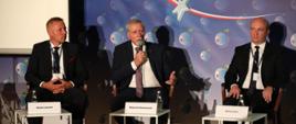 Widok na scenę i trzech panelistów: od lewej: Michał Janasik – Wiceprezes ds. Finansów i Komercjalizacji Centrum Łukasiewicz, Wojciech Kamieniecki – dyrektor NCBR, Michał Góra – Prezes Alfavox. Za panelistami plansza z logo i napisem: Economic Forum.