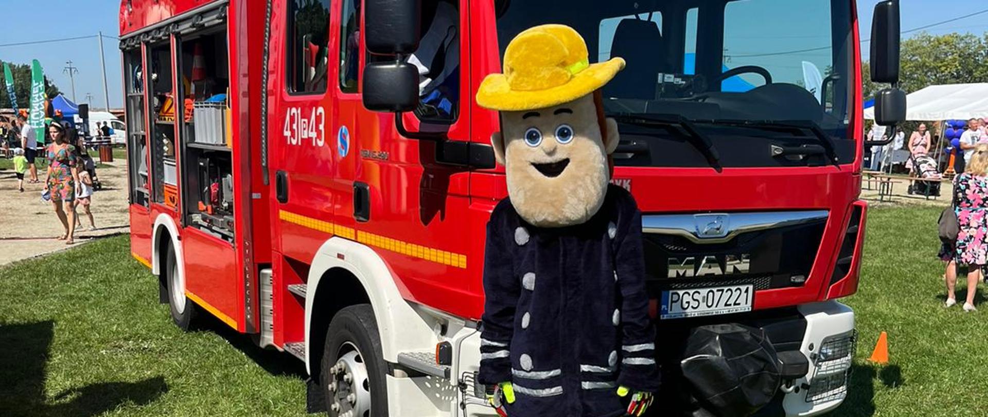 Maskotka Strażaka Sama stoi przed wozem strażackim i pozuje do zdjęcia. 