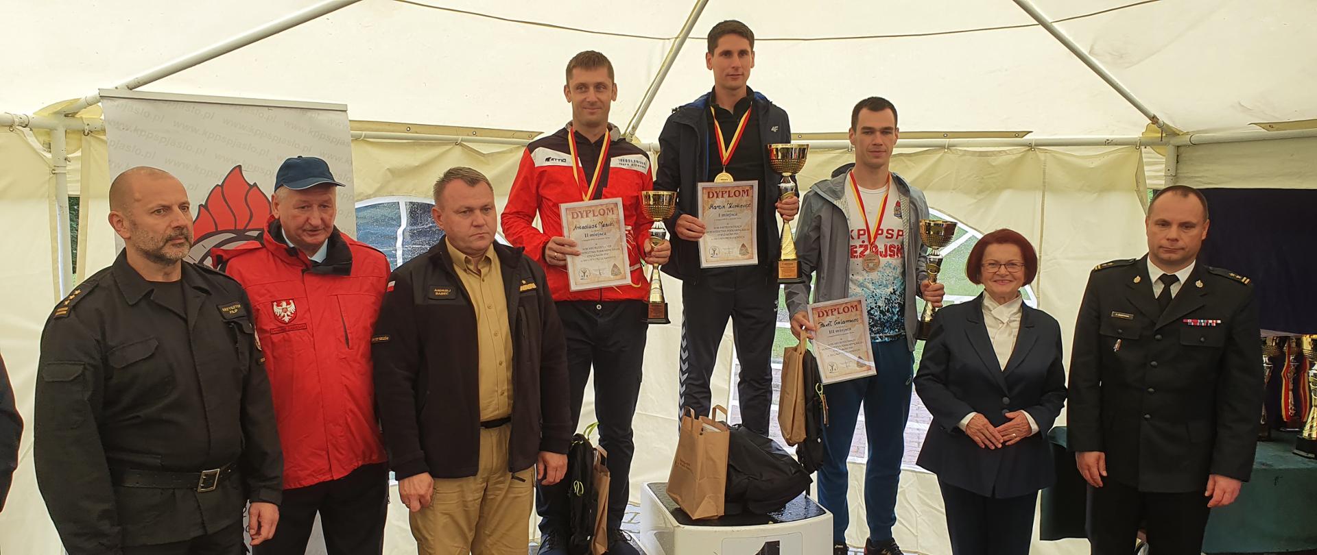 Na zdjęciu widzimy strażaków w umundurowaniu między innymi Komendanta Wojewódzkiego Państwowej Straży Pożarnej i jego zastępcę ora osoby cywilne pozujące do wspólnego zdjęcia z zwycięzcami biegu stojącymi na podium. 
