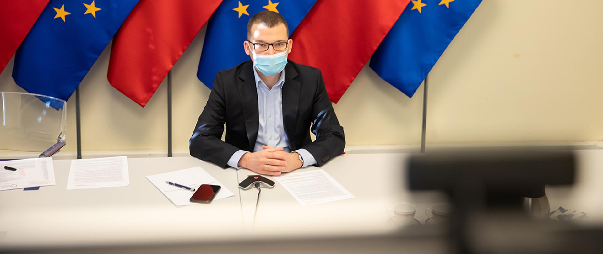 Na zdjęciu widać wiceministra Pawła Szefernakera siedzącego za stołem w maseczce na tle flag Polski i UE. Wiceminister patrzy w ekran.