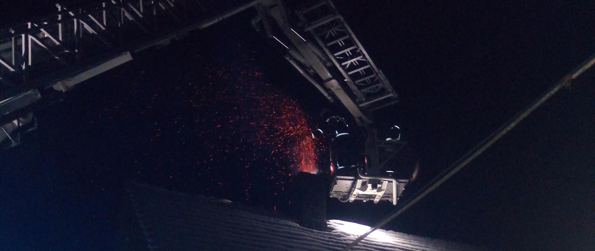 Na zdjęciu widać pojazd typu podnośnik straży pożarnej. W koszu podnośnika znajduje się dwóch strażaków którzy gaszą przewód kominowy. Z komina na dachu wydobywają się iskry.