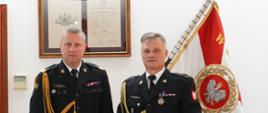 dwóch strażaków w mundurach ze sznurem stoją pod ścianą , jeden strażak trzyma w ręku medal i czerwoną teczkę, w tle Sztandar