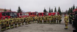 Zdjęcie wykonane na terenie zakładu Mar - rom w Radomiu po zakończonych ćwiczeniach. Na zdjęciu widać funkcjonariuszy Państwowej Straży Pożarnej jak również przedstawicieli innych podmiotów biorących udział w ćwiczeniach. Ustawieni są w rzędzie obok siebie na utwardzonym placu. Jeden z funkcjonariuszy starszy brygadier Marcin Ziomek zastępca komendanta miejskiego Państwowej Straży Pożarnej w Radomiu dokonuje omówienia i podsumowania ćwiczeń. Za plecami strażaków ustawione są pojazdy ratowniczo – gaśnicze w kolorze czerwonym z białymi i srebrnymi elementami zabudowy. 