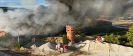 Zdjęcie przedstawia pożar obiektu przemysłowego z charakterystyczną ceglaną wieżą, wykonane z podwyższonej perspektywy. Nad miejscem zdarzenia unoszą się gęste kłęby czarnego i szarego dymu, które przesłaniają znaczną część zabudowań. W dolnej części fotografii widoczne są tory kolejowe oraz rozłożone linie gaśnicze. Na placu znajdują się samochody Państwowej Straży Pożarnej, w tym podnośnik hydrauliczny, prowadzące działania ratowniczo-gaśnicze. Wokół obiektu widoczne są pryzmy składowanego kruszywa oraz otaczające teren pola uprawne. Fotografia ukazuje dużą skalę pożaru oraz intensywne działania strażaków mające na celu opanowanie ognia i ograniczenie rozprzestrzeniania się dymu.