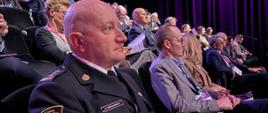 Strażak w mundurze wyjściowym, w pozycji siedzącej na sali widowiskowej. Za nim kilkadziesiąt innych uczestników wydarzenia.