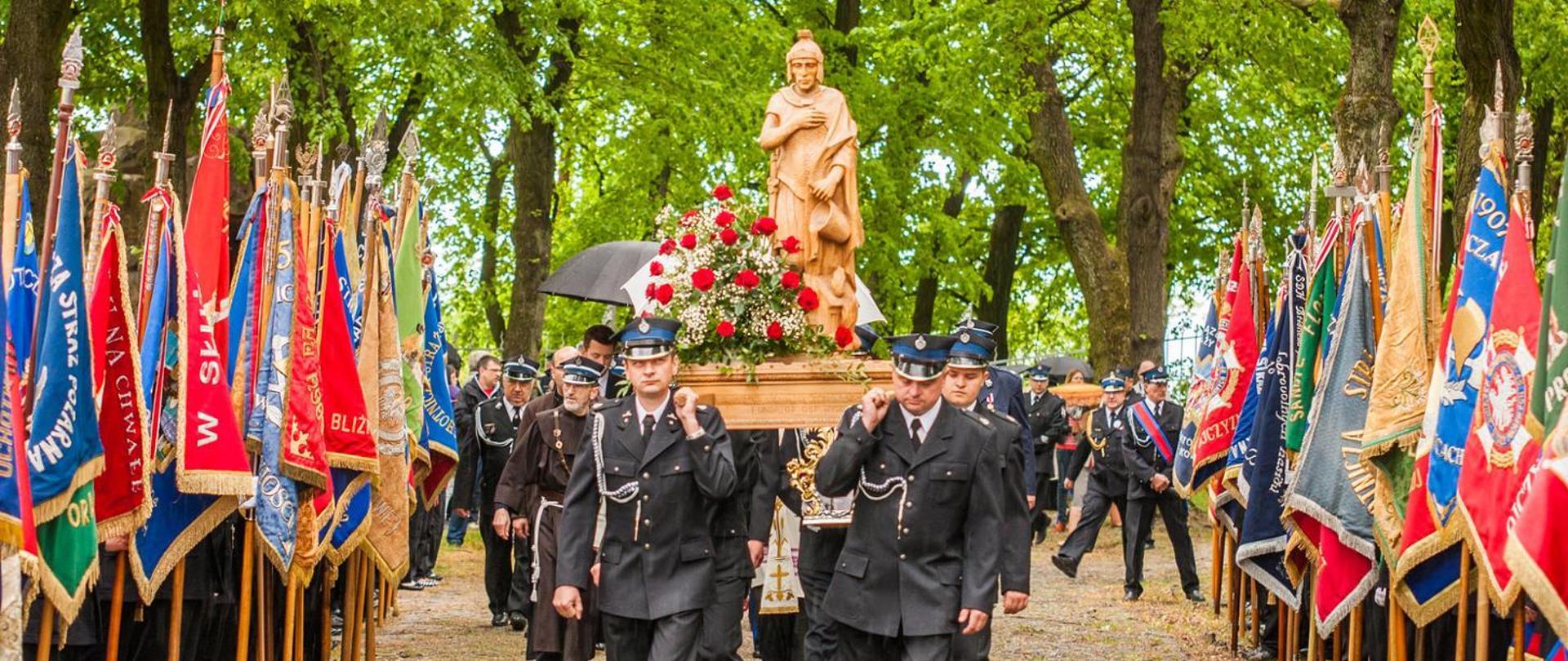 Uroczystości w ramach pielgrzymki strażaków do Sanktuarium Świętej Anny Samotrzeciej na Górze Świętej Anny. Strażacy ochotnicy w mundurach galowych niosą na barkach drewnianą figurę Świętego Floriana. Po lewej i prawej stronie widoczne poczty sztandarowe OSP. W tle widoczne drzewa.