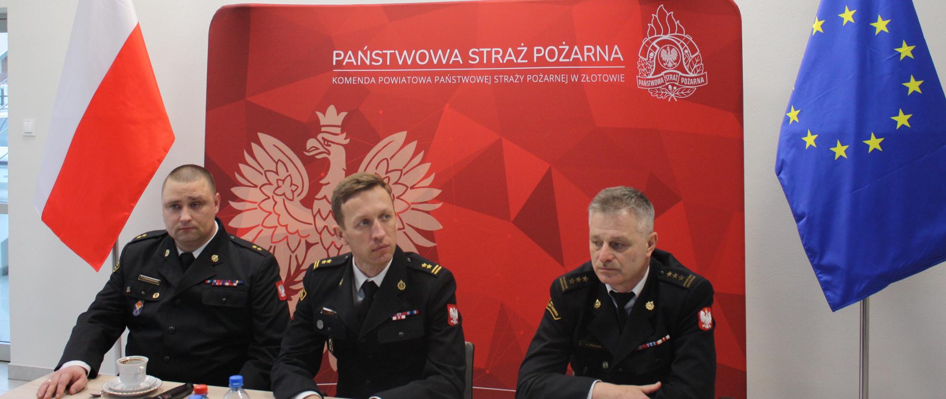 trzech strażaków - komendantów siedzi przy stole, za nimi ścianka tekstylna z czerwonym tłem , logo PSP i napisem państwowa straż pożarna, z lewej strony ścianki stoi biało czerwona flaga, z prawej strony niebieska z gwiazdkami , flaga unii europejskiej